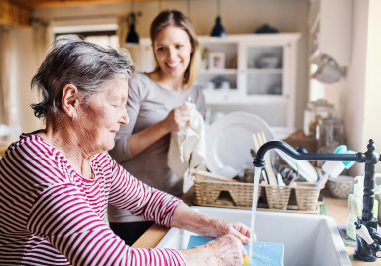 An older woman washing dishes with the help of her support worker.