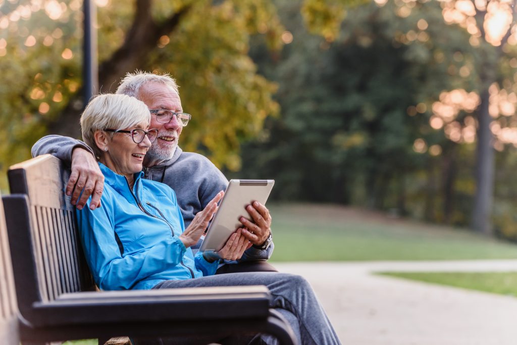 An older couple sitting on a bench looking at a tablet.