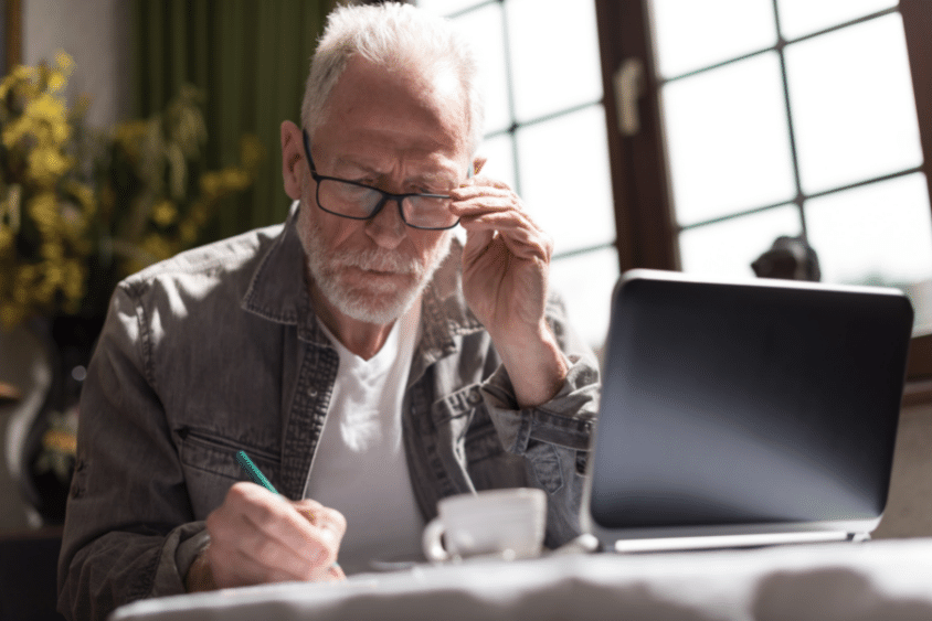 Older man writing on document and working on laptop at home