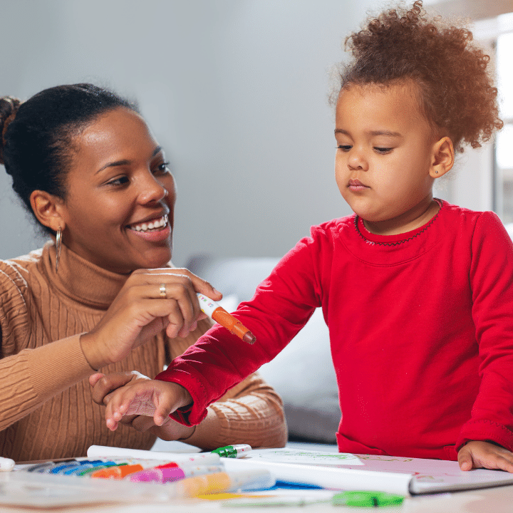 young child reaching for colourful markers with support worker in living room environment