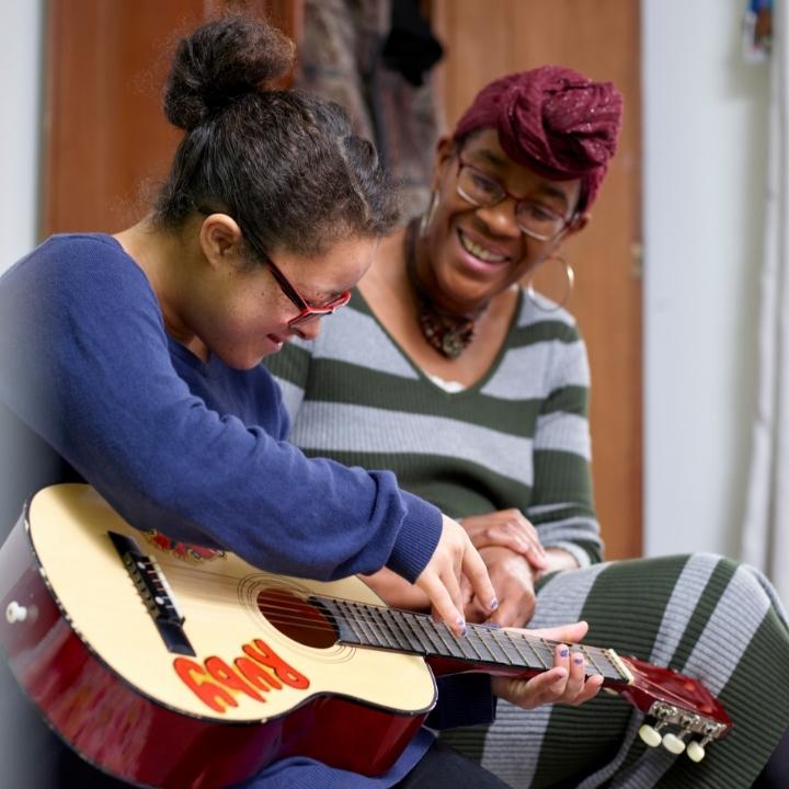 Two women pictured sitting together. Young woman is playing musical instrument.