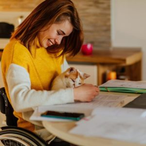 A woman in a wheelchair studying at home with her cat in her lap.