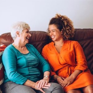 woman in orange dress smiling next to older woman in blue shirt sitting on living room couch