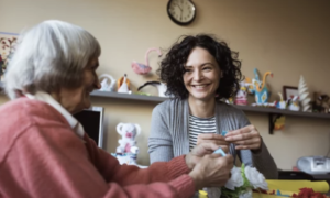 An older woman and her support worker sit knitting together.