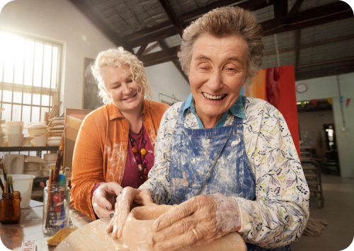 Two people are turning clay while smiling in a studio. There is a client throwing clay at the front. There is an aged care independent support worker watching from the back. There is sunlight coming through a studio window.