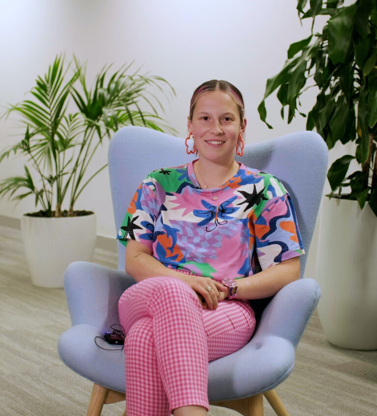 Annie sitting on a chair facing camera wearing a floral shirt and smiling.