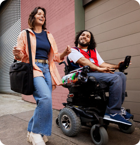 Two people are smiling as they move past buildings in a street. The client is in an electric wheelchair. The disability worker is beside them.