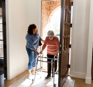 An older client returns home with their aged care independent support worker. The client uses a walking frame through their open front door. The worker helps the client to walk.