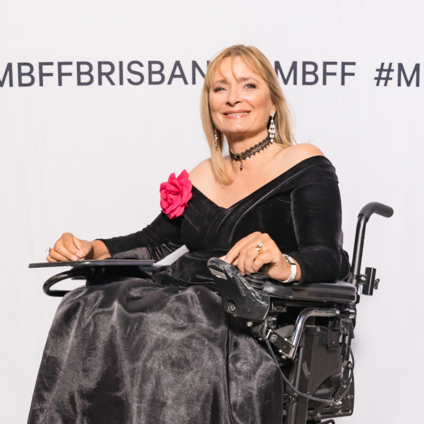 Carol Taylor poses for a photo at an event. She's wearing a black gown with a pink flower pinned on her right shoulder. She has straight blonde hair that sits just past her shoulders.