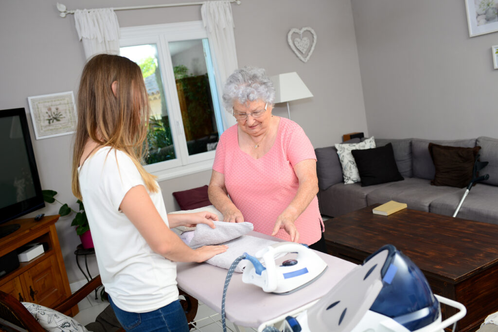 cheerful young woman ironing and helping with household chores for an elderly woman at home