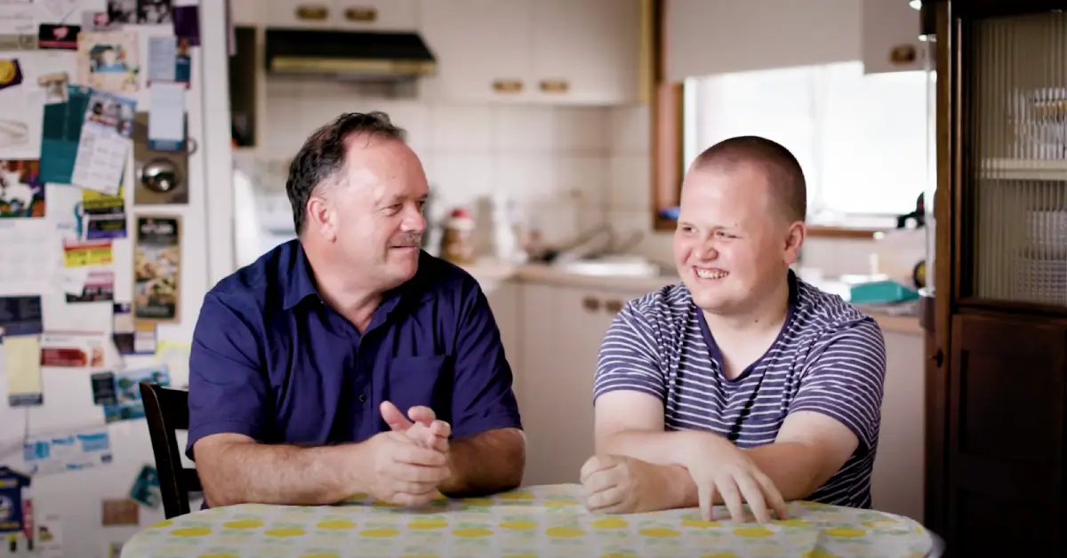 Liam and his support worker Brian sit smiling at a kitchen table.