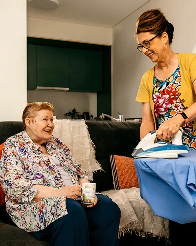 Older person with a walking stick sits on the couch with tea and chats to support worker ironing.