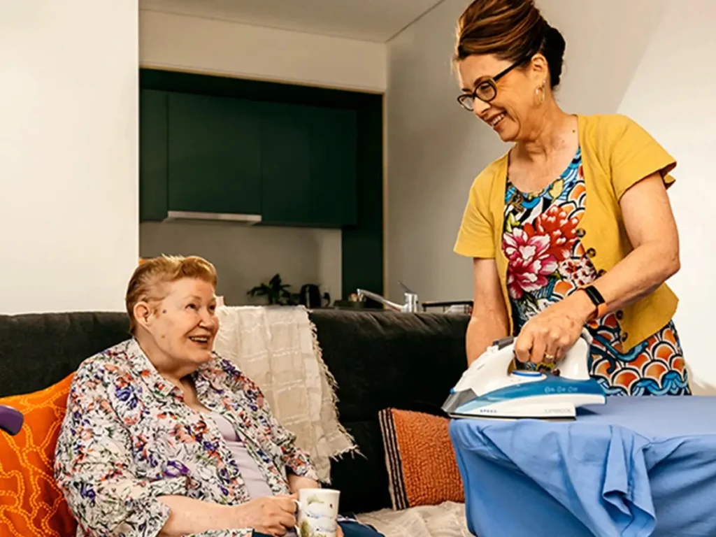 Older person with a walking stick sits on the couch with tea and chats to support worker ironing.