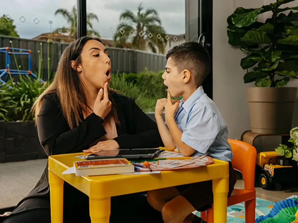 Speech pathologist and child with speech disorder point to their chins to make O shape with mouths.