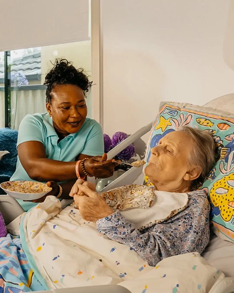 Support worker smiles as they feed an older person living with Dementia laying in bed at home.