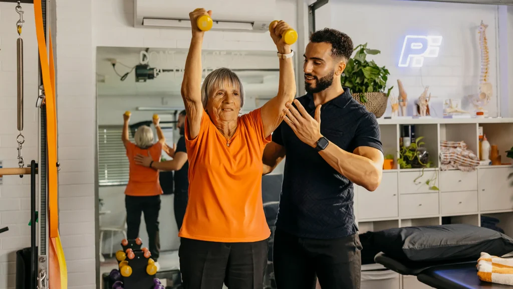 Physiotherapist supports older client’s arm and back as they lift light dumbbells above their head.