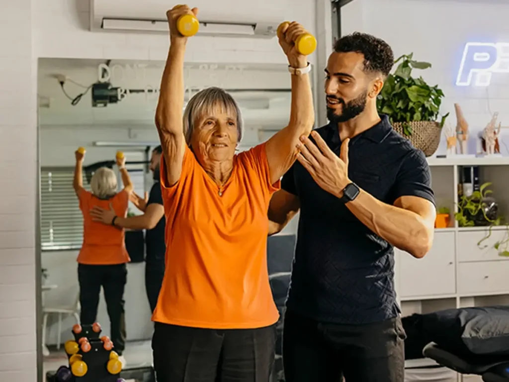 Physiotherapist supports older client’s arm and back as they lift light dumbbells above their head.