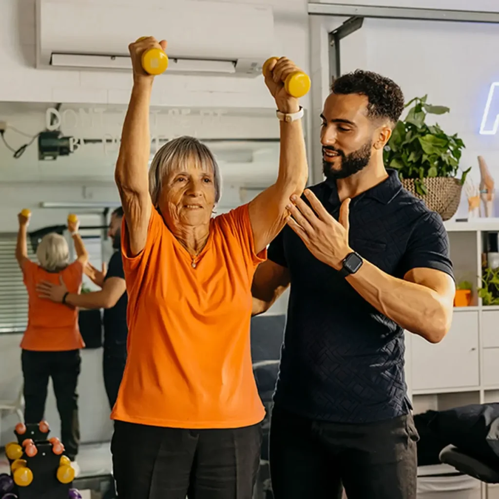 Physiotherapist supports older client’s arm and back as they lift light dumbbells above their head.