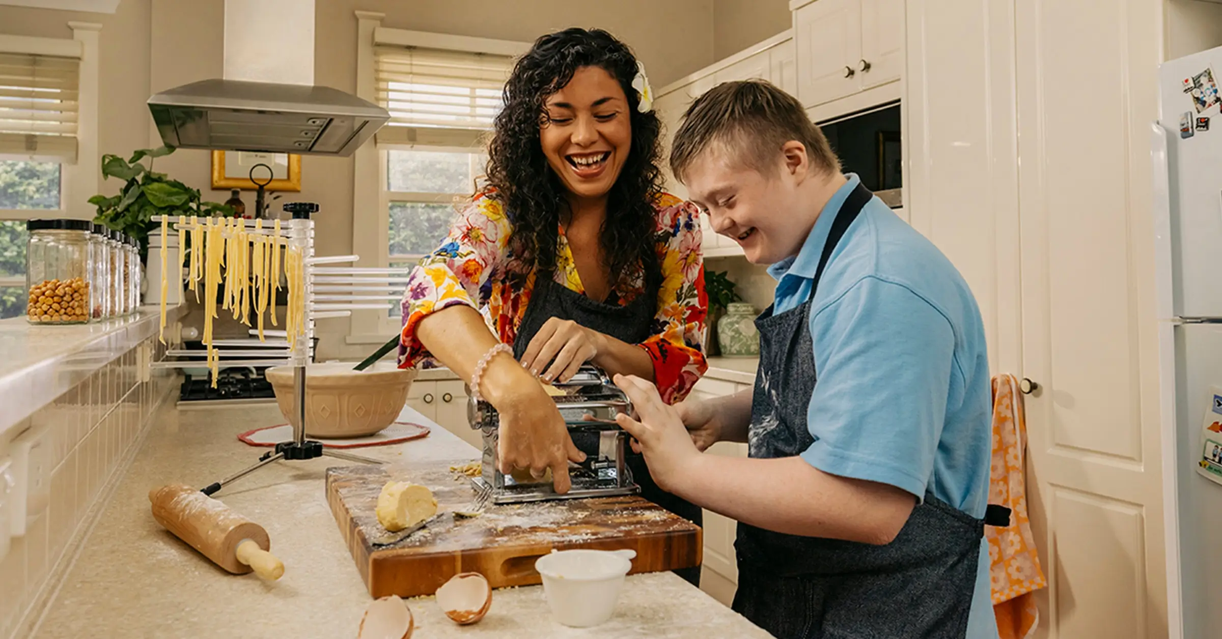Support worker and child with disability in aprons roll pasta through a dough machine in kitchen.