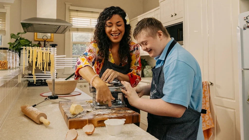 Support worker and child with disability in aprons roll pasta through a dough machine in kitchen.