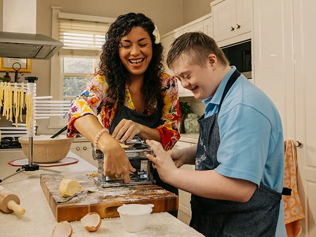 Support worker and child with disability in aprons roll pasta through a dough machine in kitchen.