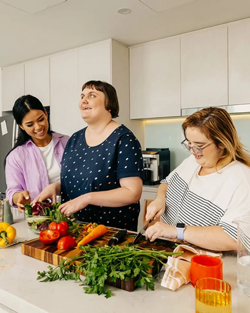 Support worker helps two people with disabilities prepare vegetables at the kitchen bench.