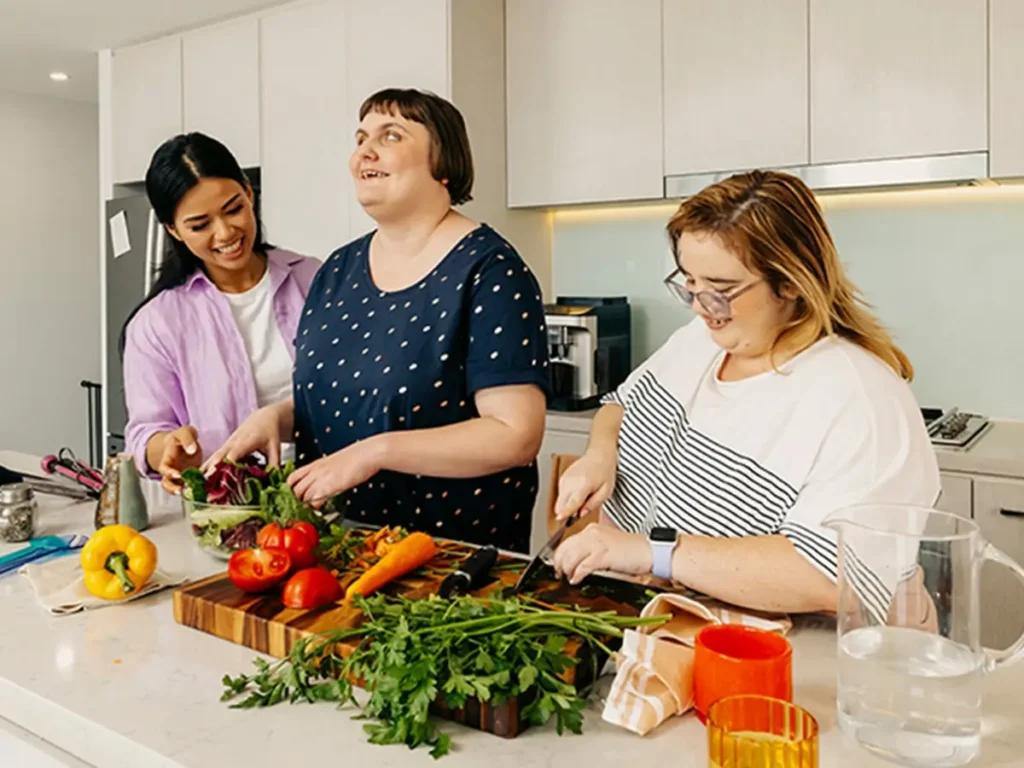 Support worker helps two people with disabilities prepare vegetables at the kitchen bench.