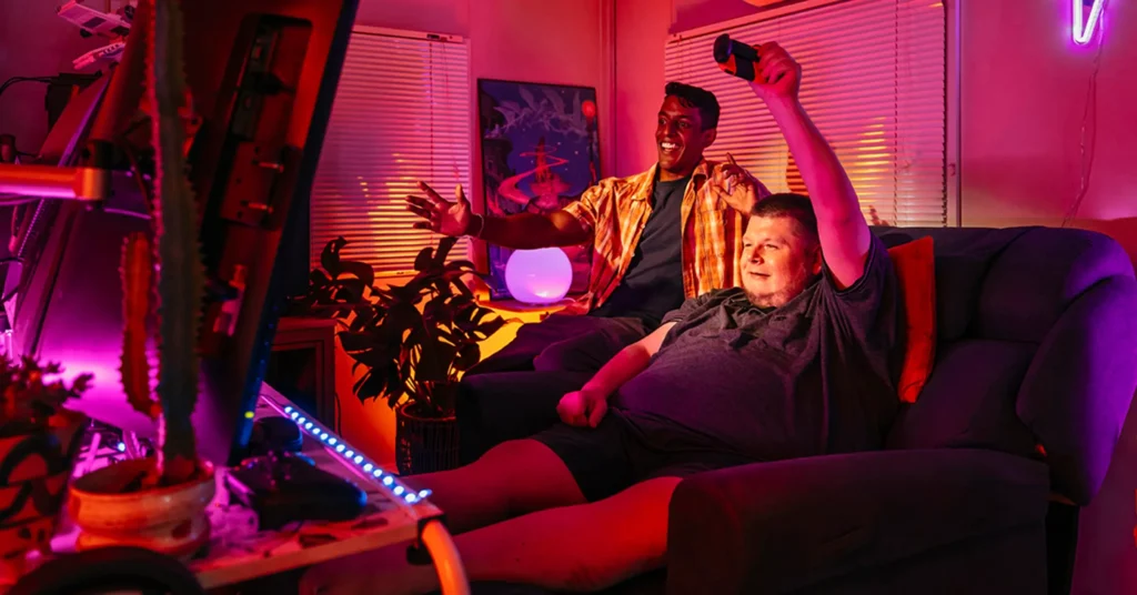 Support worker and person with disability celebrate video game win in a pink neon-lit gamer room.