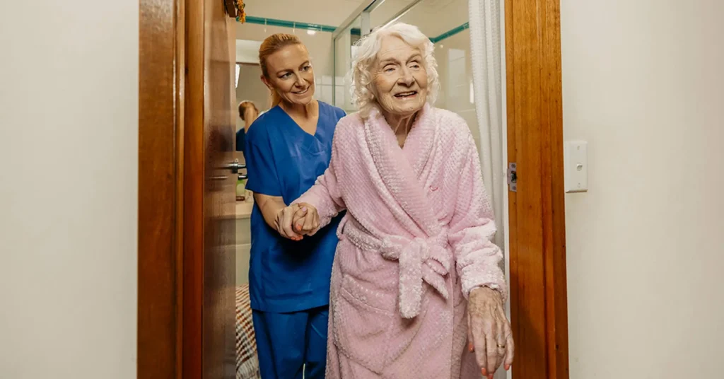 Registered Nurse in scrubs helps older client in pink bathrobe walk out of their bathroom.