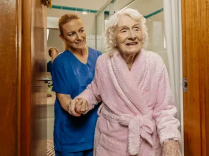 Registered Nurse in scrubs helps older client in pink bathrobe walk out of their bathroom.