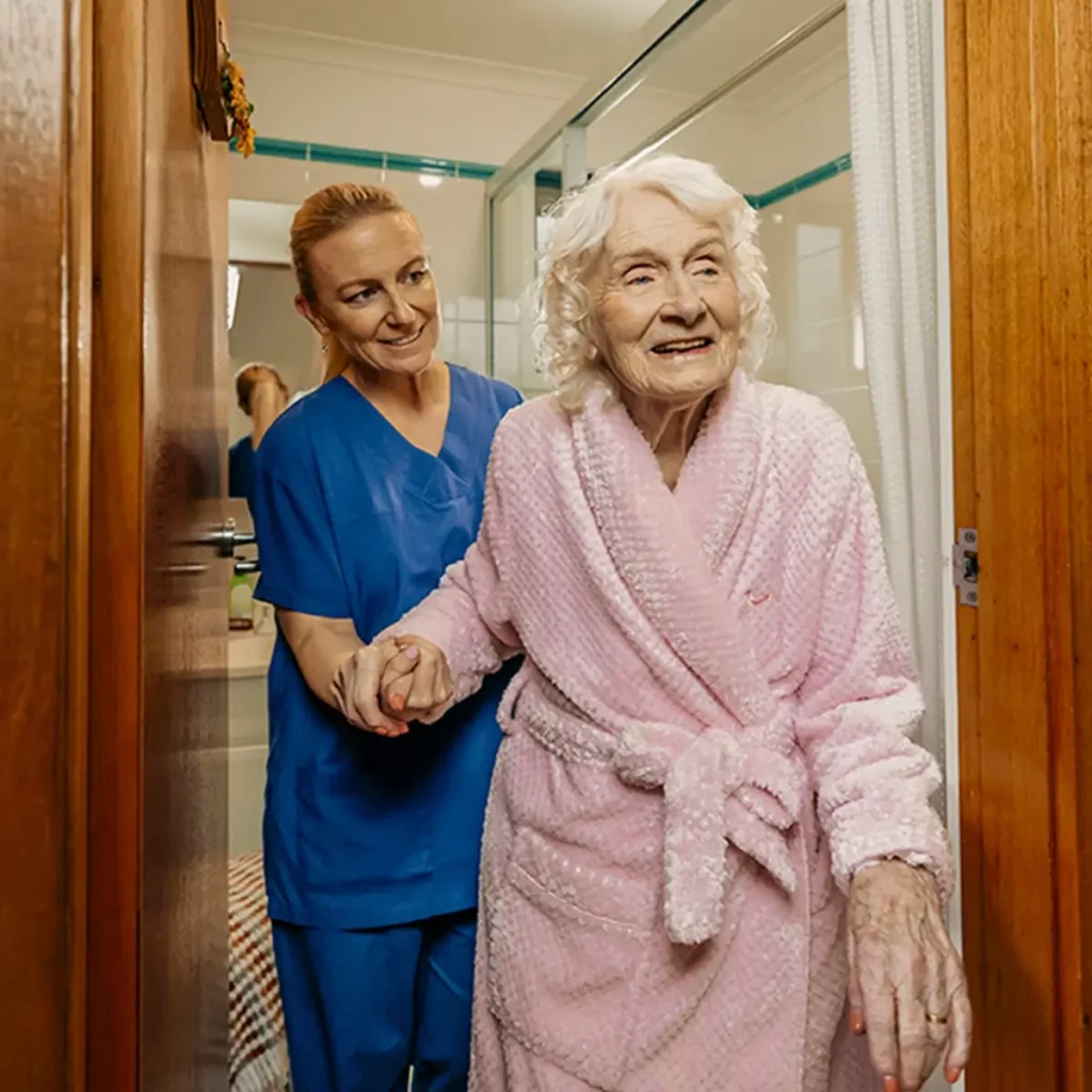 Registered Nurse in scrubs helps older client in pink bathrobe walk out of their bathroom.