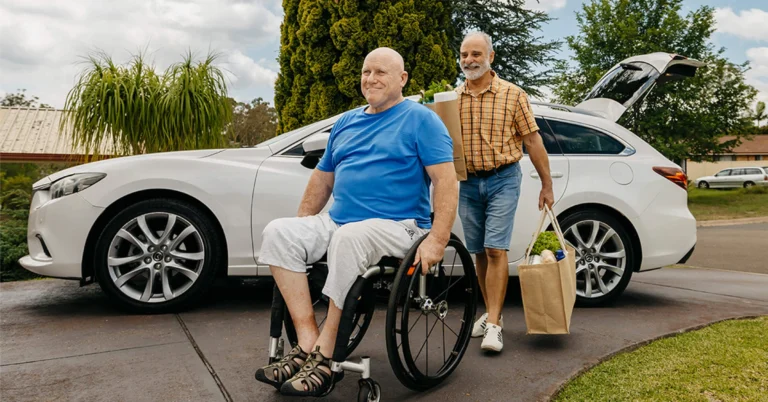 Person who is blind and paraplegic wheels up driveway with support worker following with groceries.