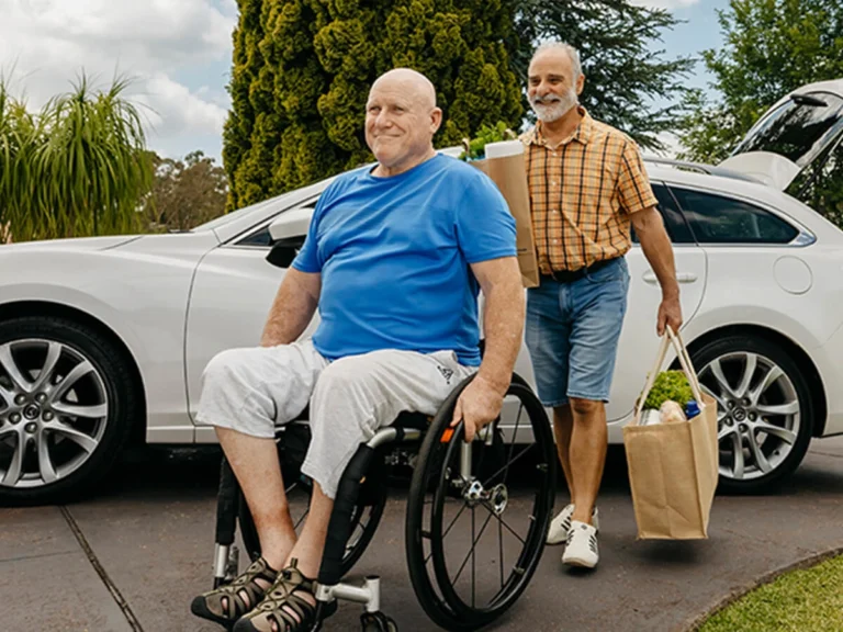 Person who is blind and paraplegic wheels up driveway with support worker following with groceries.