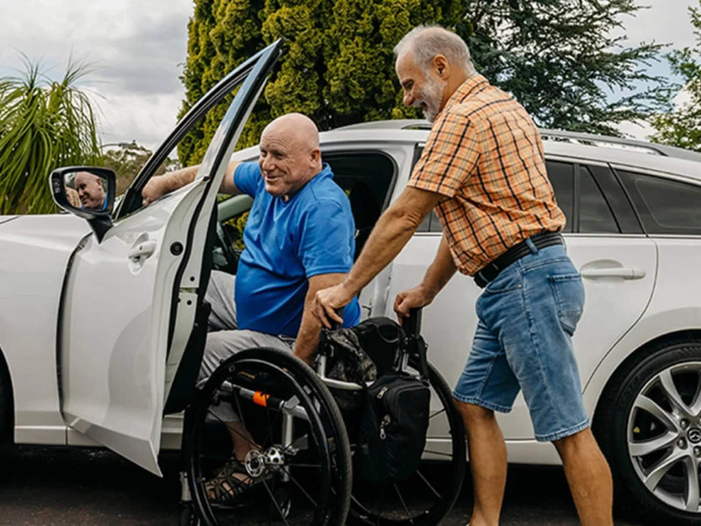 Older support worker holds wheelchair at car door for client who is blind and paraplegic.