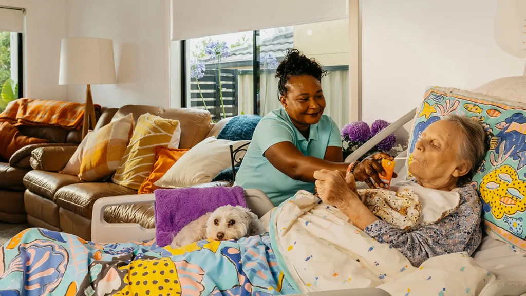 Support worker smiles as they feed an older person living with Dementia laying in bed at home.