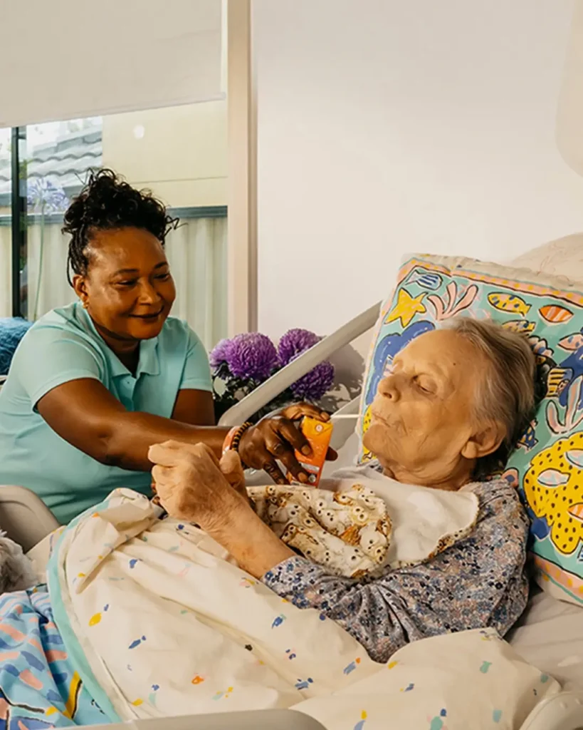 Support worker smiles as they feed an older person living with Dementia laying in bed at home.
