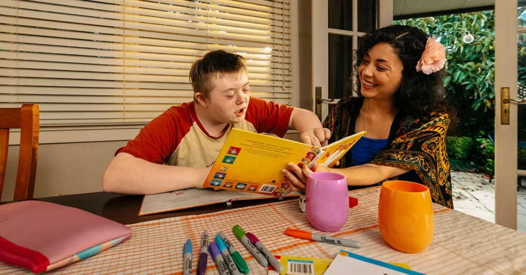 Support worker helps child with disability finger-point read picture book at the dining table.