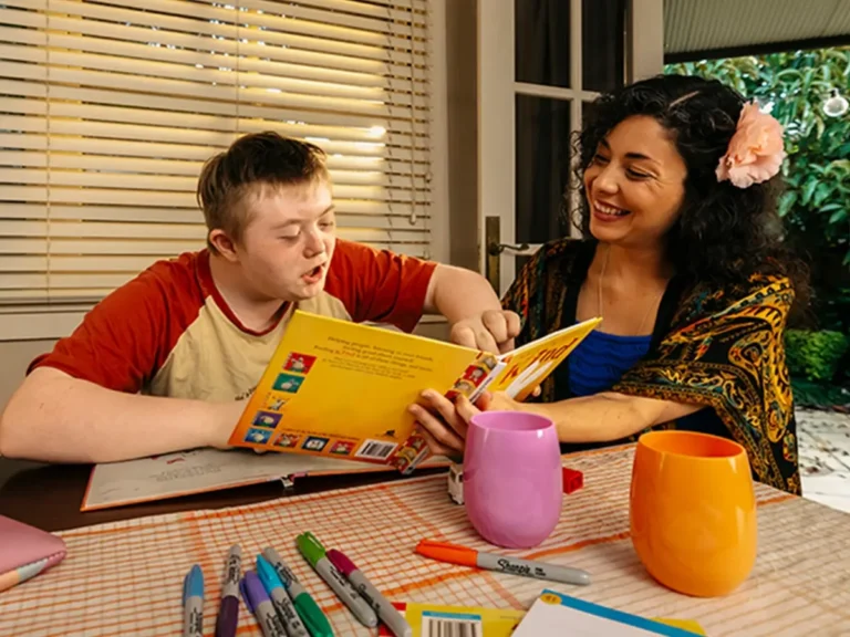 Support worker helps child with disability finger-point read picture book at the dining table.