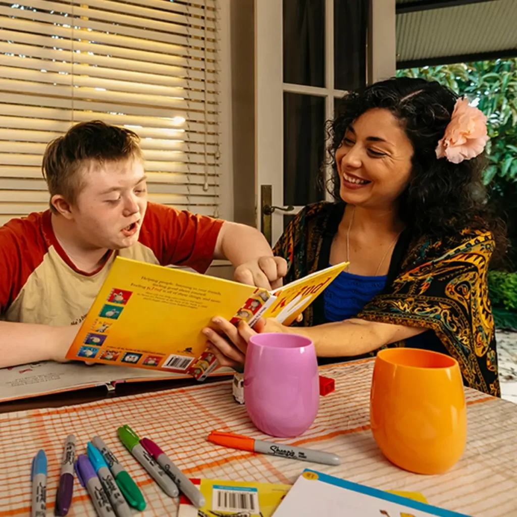 Support worker helps child with disability finger-point read picture book at the dining table.