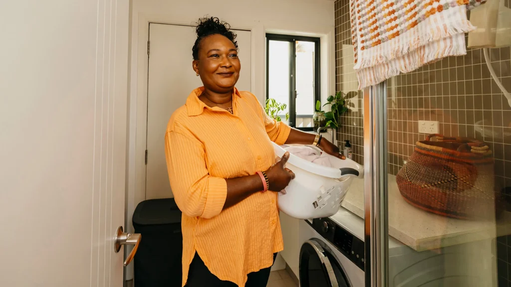 Support worker carries a washing basket on hip through the laundry at their client’s home.