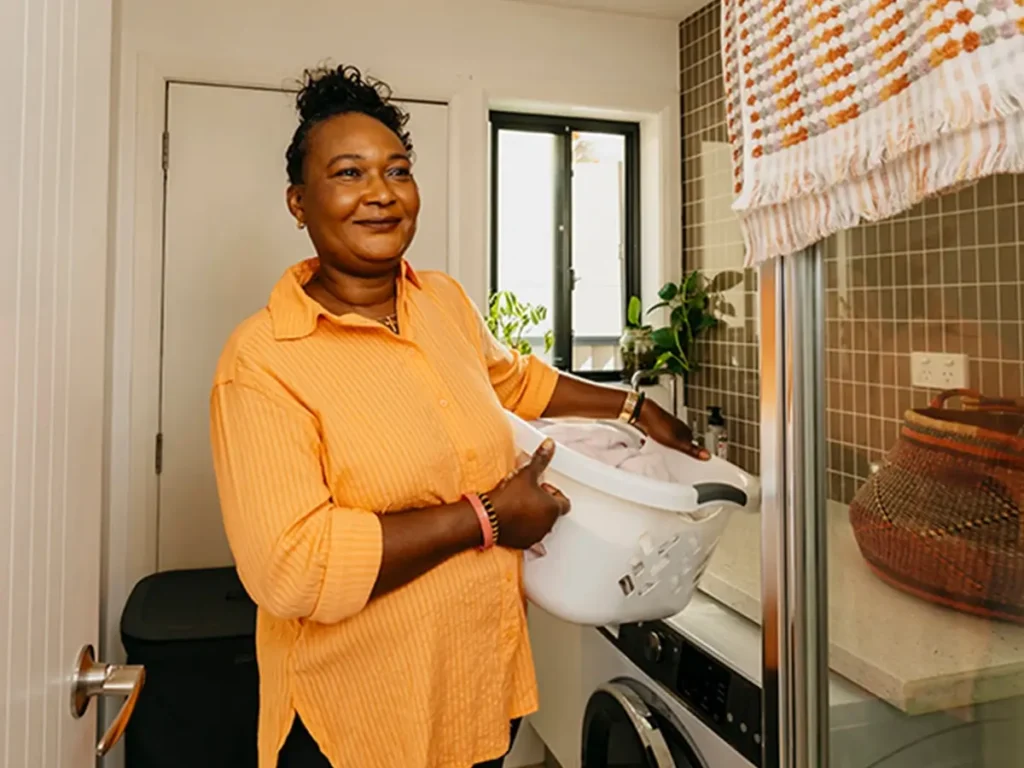 Support worker carries a washing basket on hip through the laundry at their client’s home.