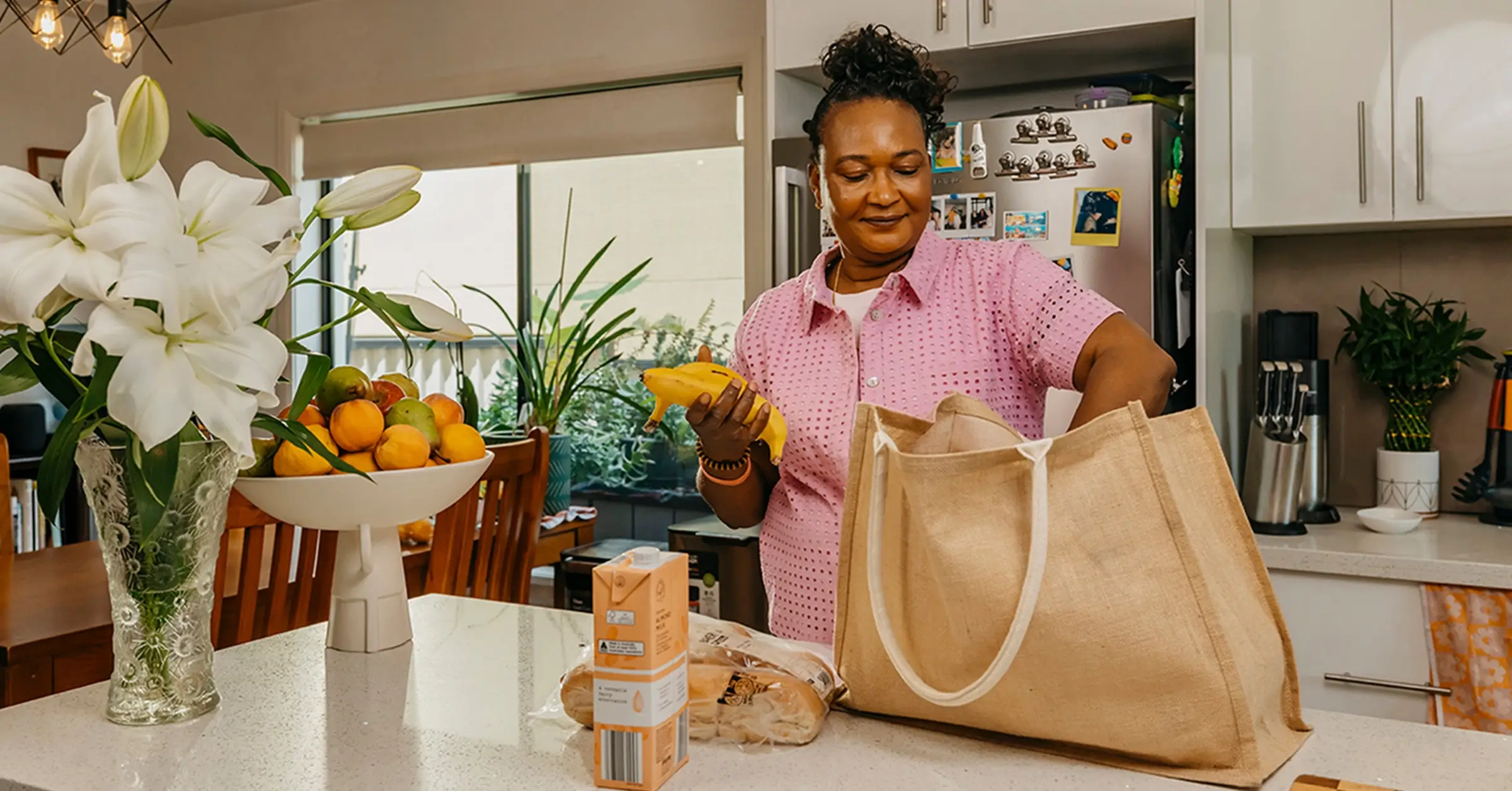 Support worker unpacks groceries from a jute shopping bag at their client’s kitchen bench.