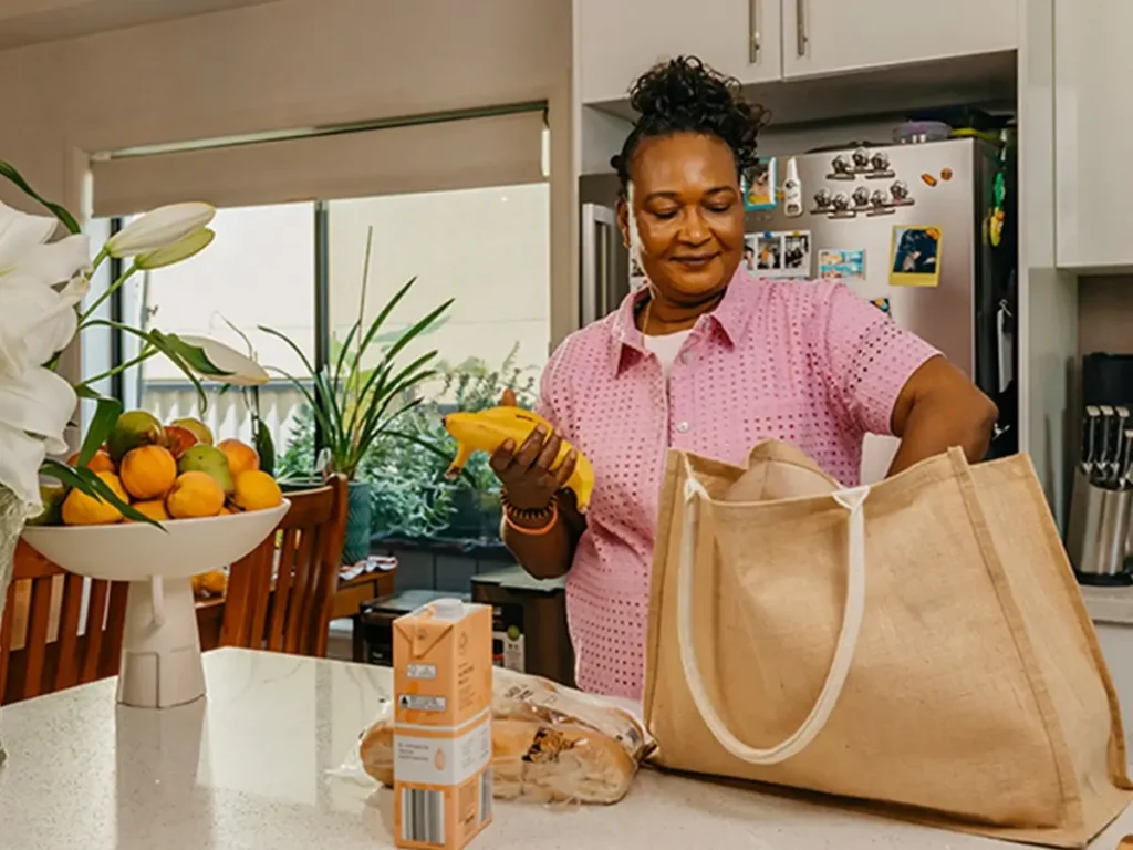 Support worker unpacks groceries from a jute shopping bag at their client’s kitchen bench.