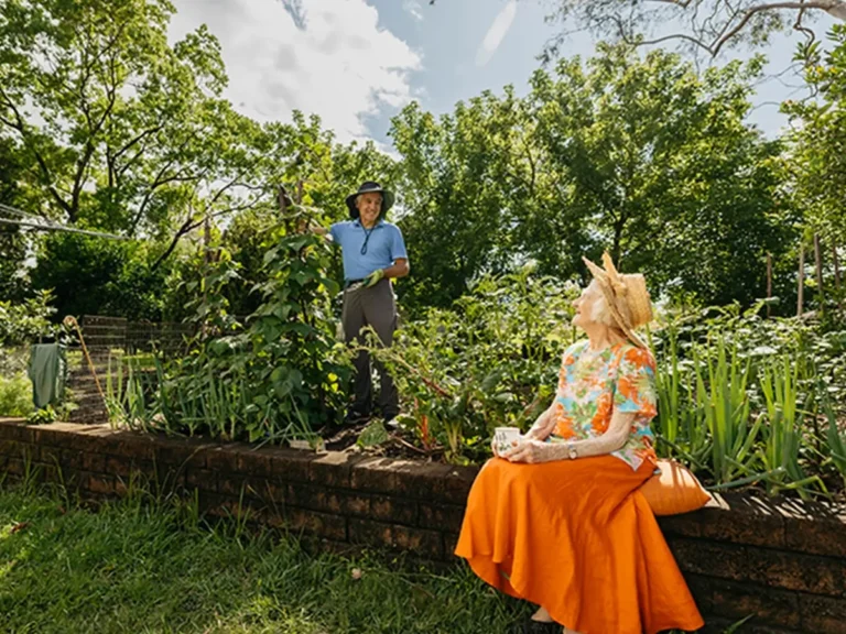 Support worker prunes plant in garden while chatting to their older client sitting on garden wall.