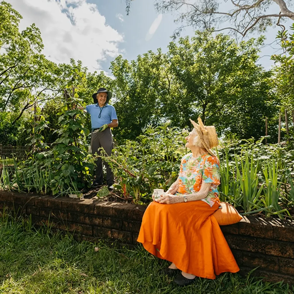 Support worker prunes plant in garden while chatting to their older client sitting on garden wall.