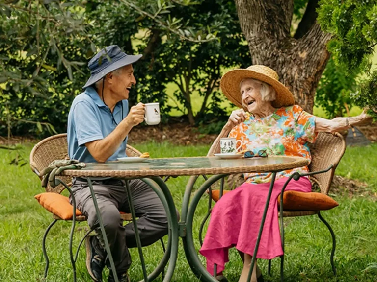 Support worker prunes plant in garden while chatting to their older client sitting on garden wall.