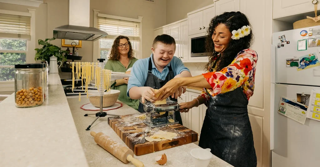 Mum watches support worker and her child with disability roll pasta through dough machine. 