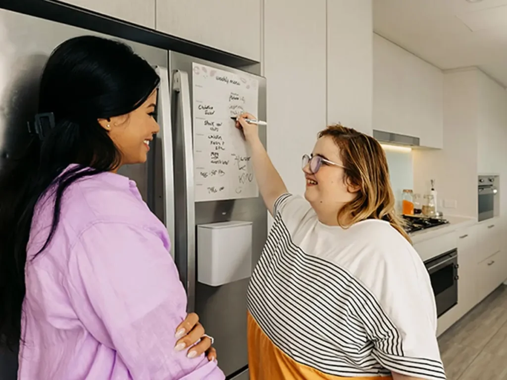 Person with disability in kitchen writes list on fridge while support worker watches.