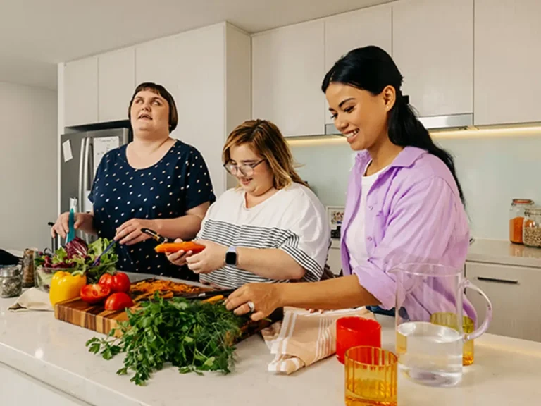 Support worker helps two people with disabilities prepare vegetables at the kitchen bench.