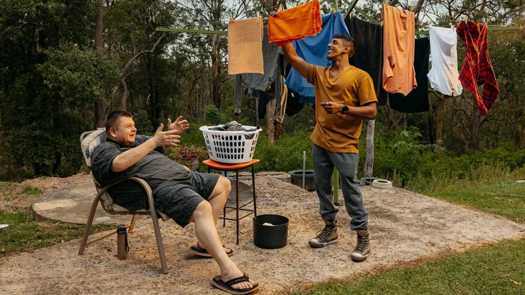 Support worker hangs washing on Hills Hoist clothesline while their client with disability talks.